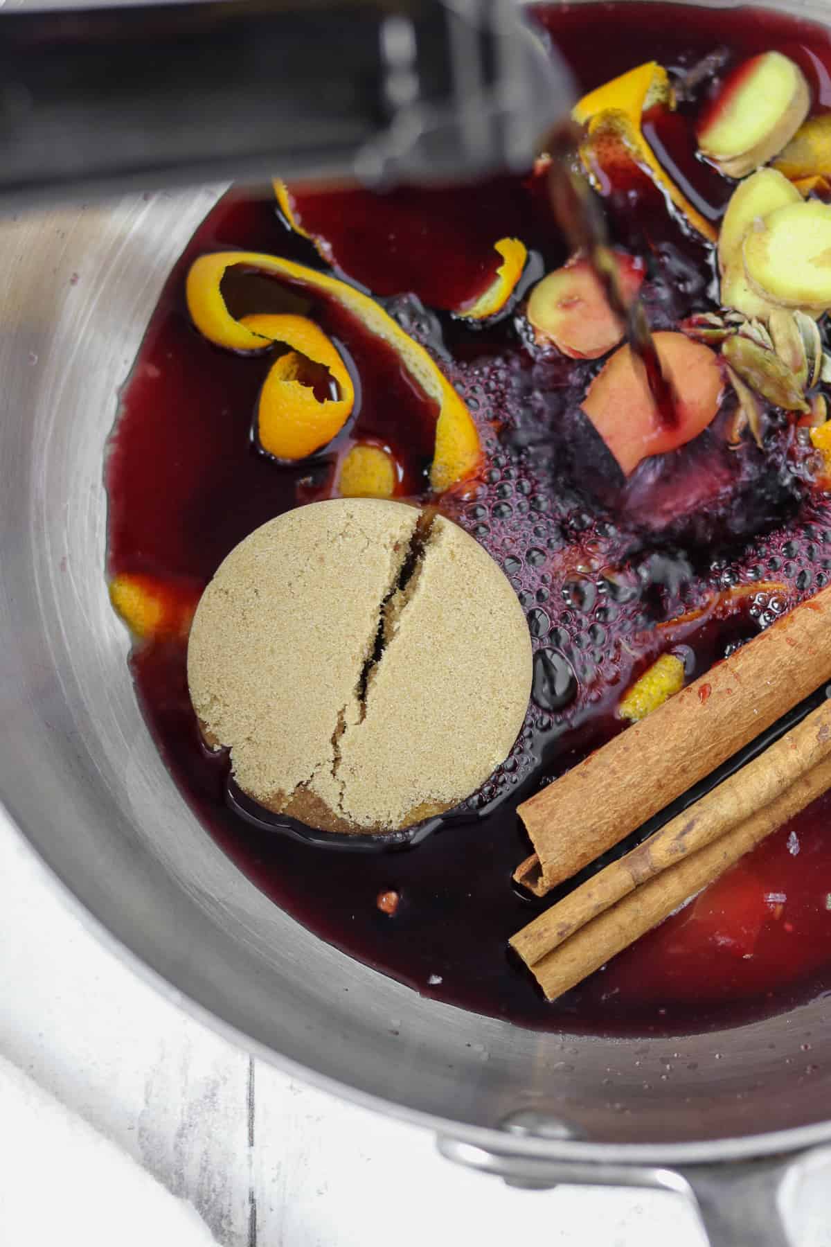 Someone pouring red wine into a pan with brown sugar, spices and orange.