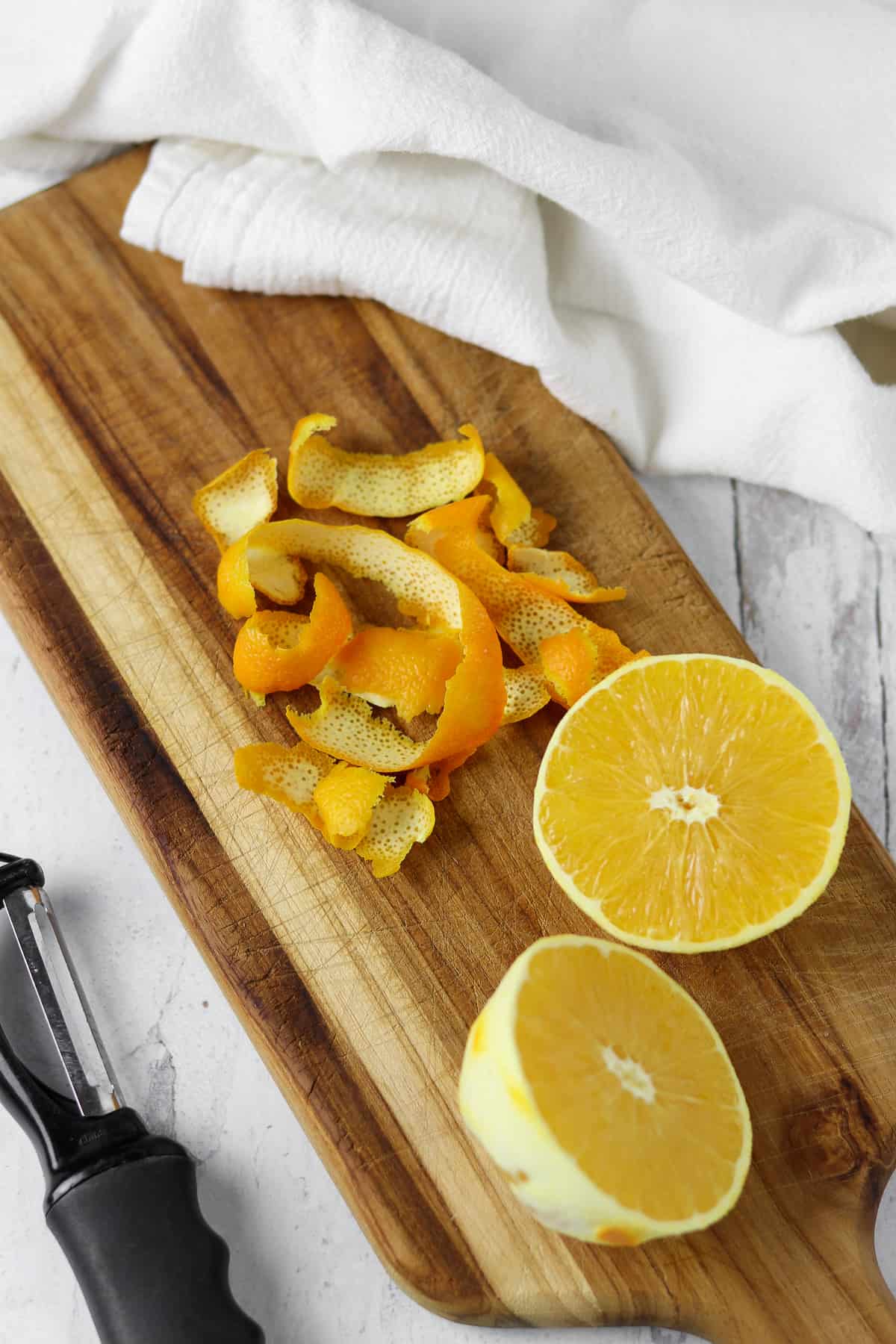 An orange, peeled and cut in half on a wooden cutting board next to a peeler and a towel.