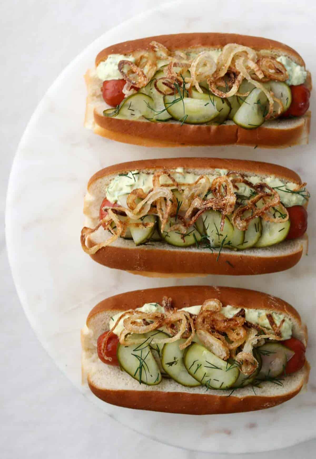 Three hot dogs topped with cucumbers and shallots on a marble surface.