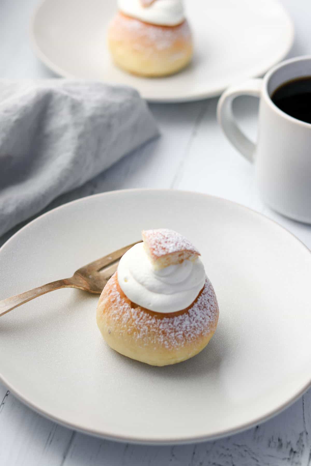 Semlor bun a on a plate next to a fork, napkin and cup of coffee.