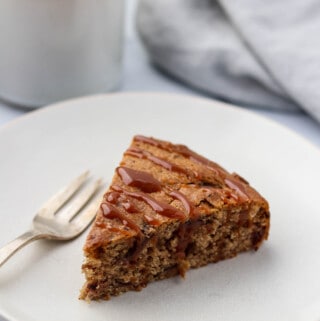 A slice of date cake drizzled with caramel on a plate next to a fork and cup of coffee.