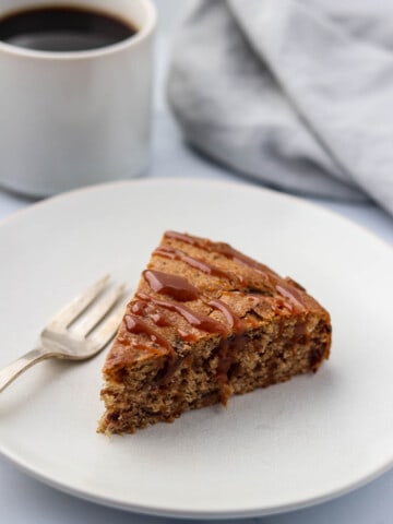 A slice of date cake drizzled with caramel on a plate next to a fork and cup of coffee.