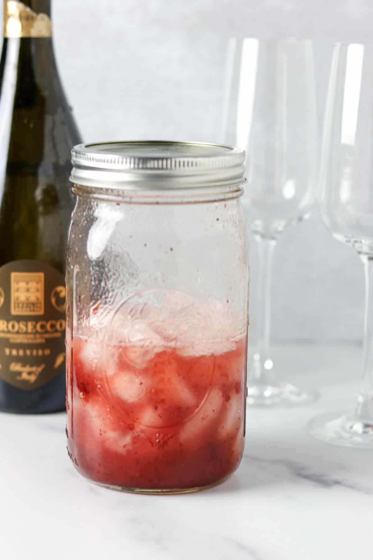 Mason jar filled with ice and a cocktail mixture next to champagne glasses and a bottle of Prosecco.