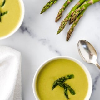Two bowls of Creamy Asparagus, Leek and Pea Soup next to asparagus spears and a spoon.