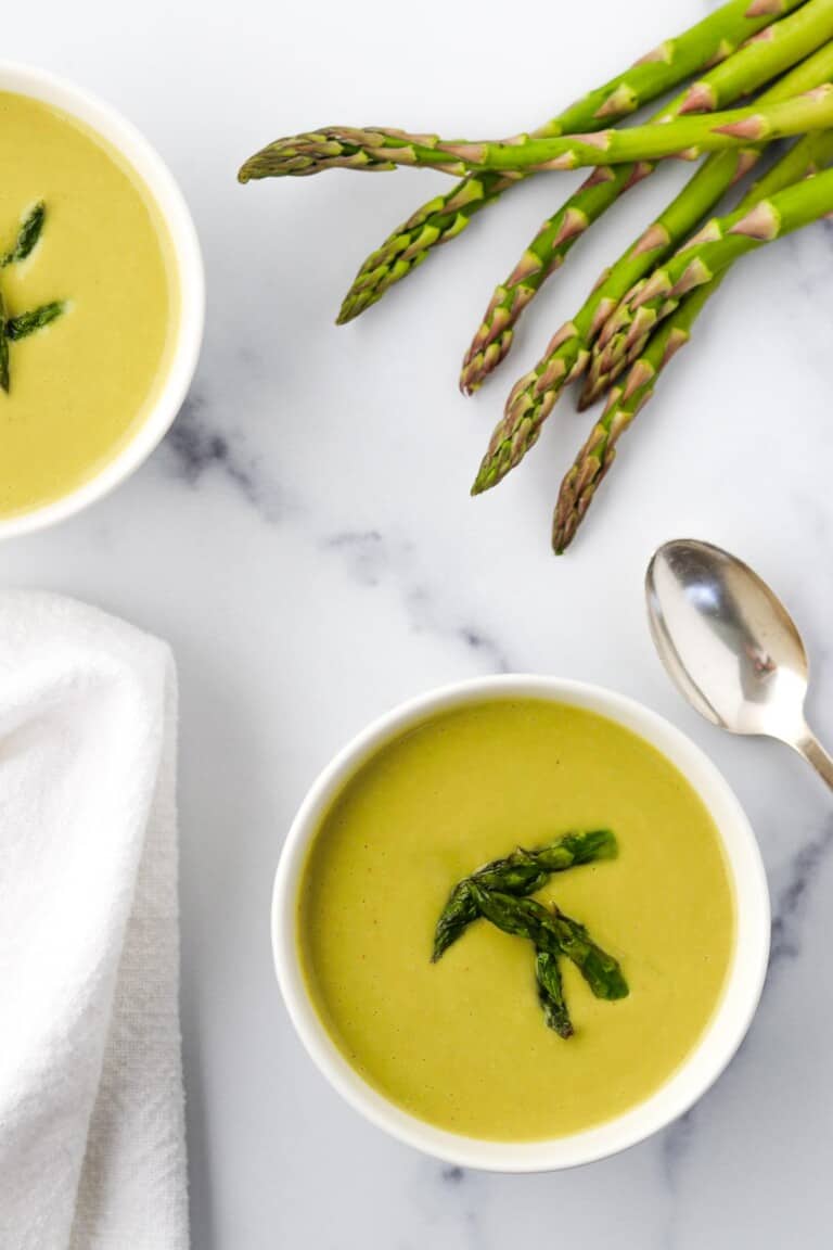 Two bowls of Creamy Asparagus, Leek and Pea Soup next to asparagus spears and a spoon.