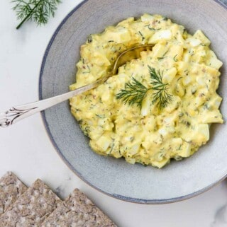 Egg salad in a bowl with a spoon next to crackers and dill sprigs.