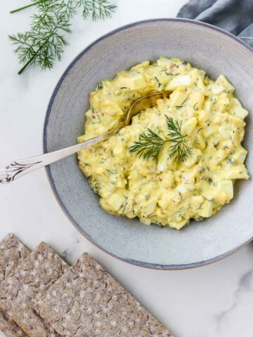 Egg salad in a bowl with a spoon next to crackers and dill sprigs.