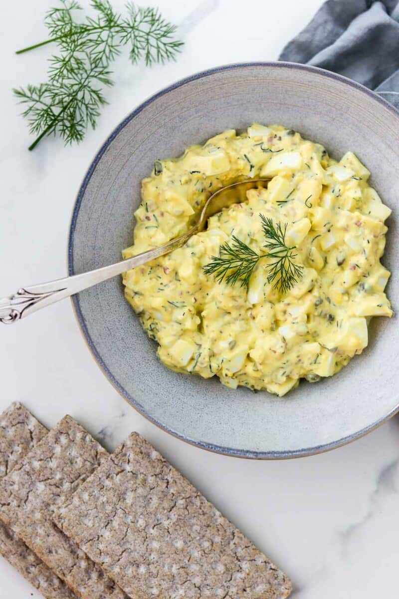 Egg salad in a bowl with a spoon next to crackers and dill sprigs.