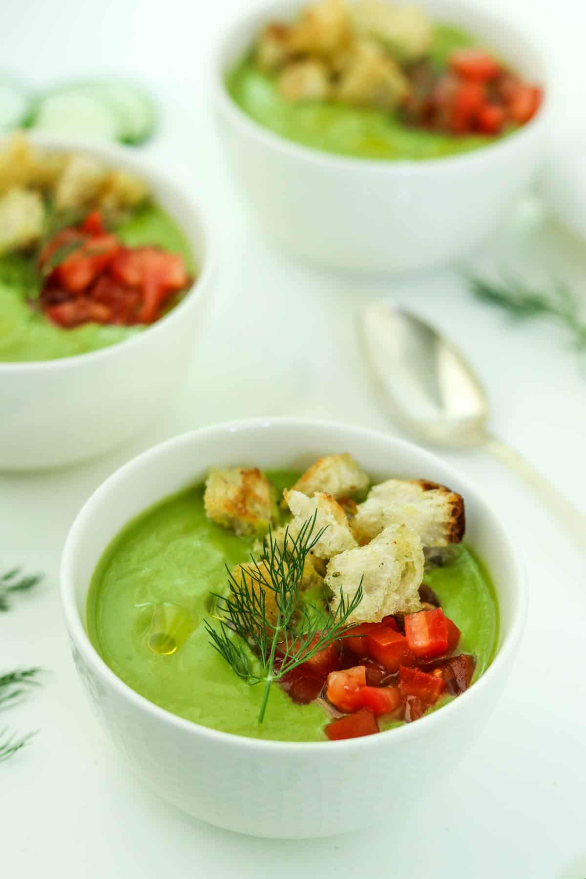 Three bowls of chilled cucumber soup topped with croutons, dill and chopped tomatoes.