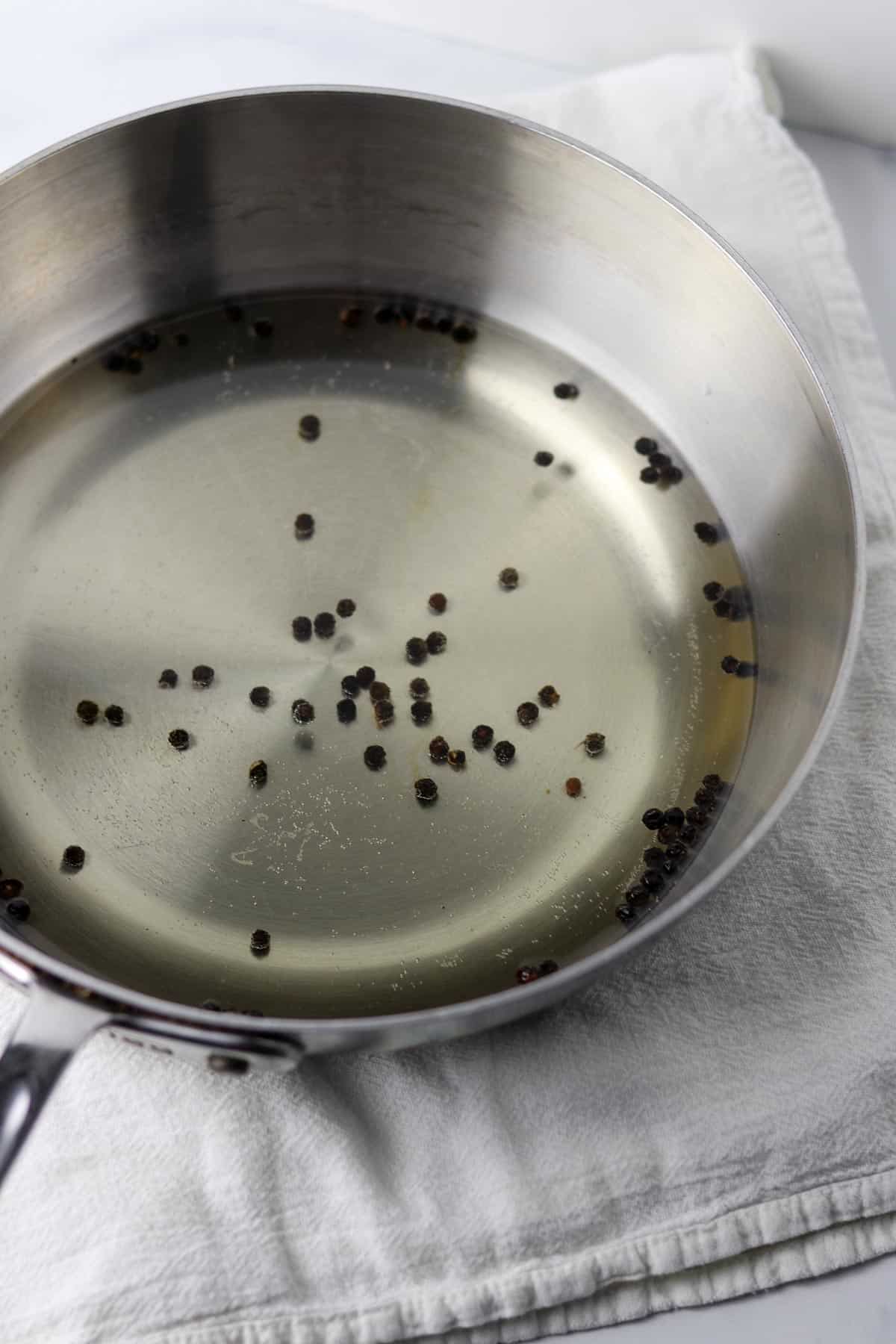 A saucepan filled with water and whole peppercorns on a white kitchen towel.