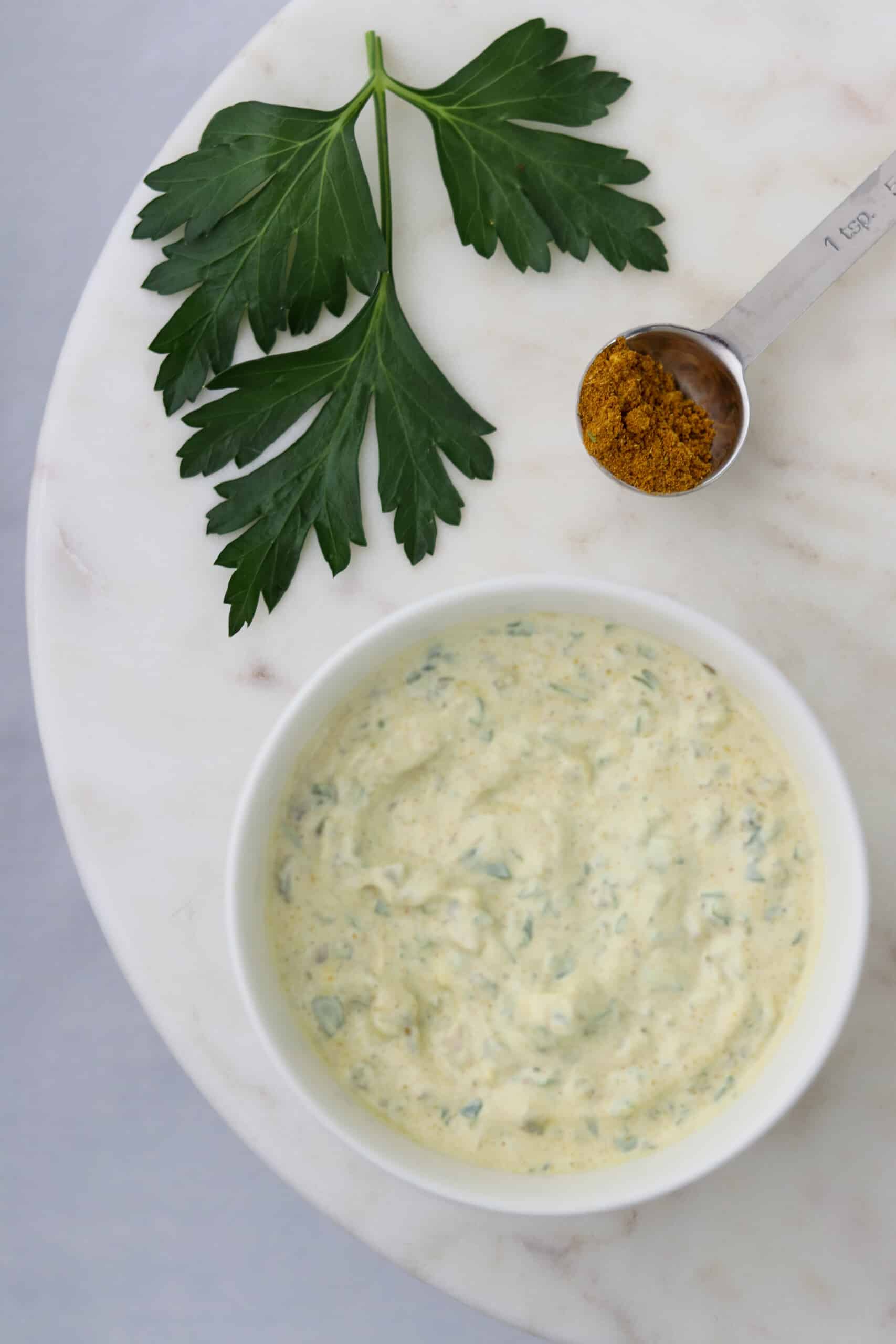 A bowl of Danish Remoulade sauce next to a sprig of parsley and curry powder.