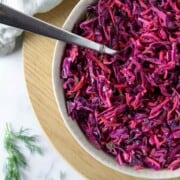 Red cabbage slaw in a bowl with a spoon on a wooden surface.