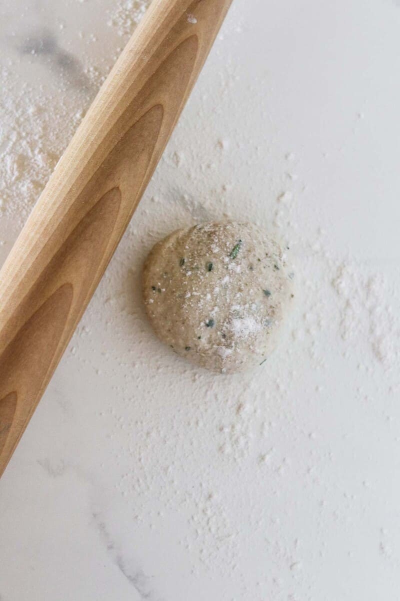 Flatbread dough on a marble surface dusted with flour next to a rolling pin.