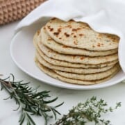 Stack of flatbreads on a plate next to herbs and a spiked rolling pin.