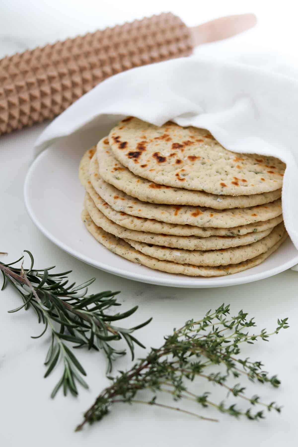Stack of flatbreads on a plate next to herbs and a spiked rolling pin.