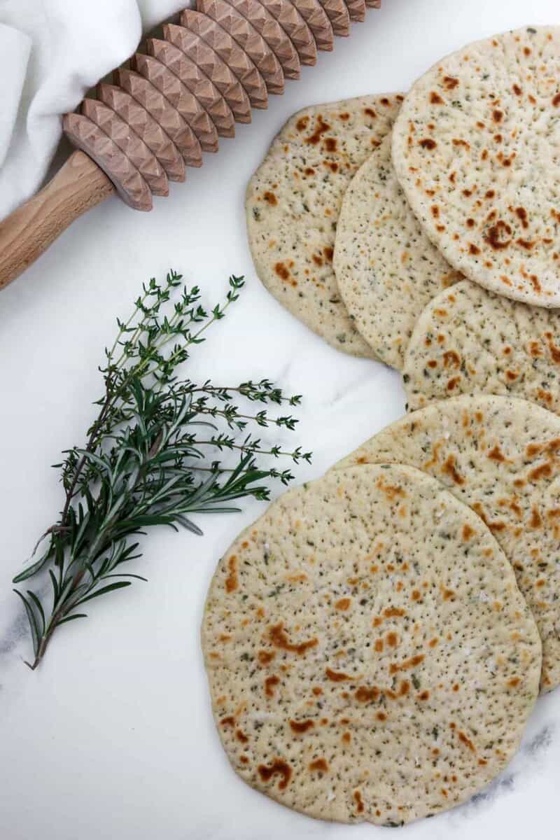 Flatbreads on a marble surface next to herbs and a spiked rolling pin.