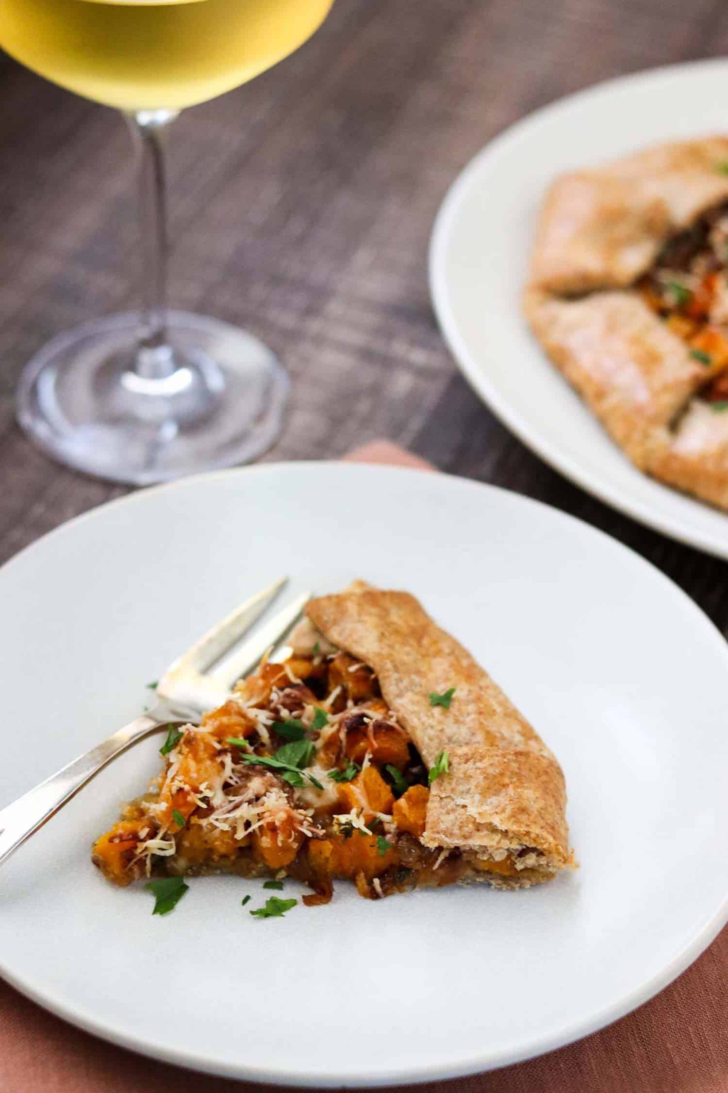 Slice of Butternut Squash Galette on a white plate with a fork and white wine.