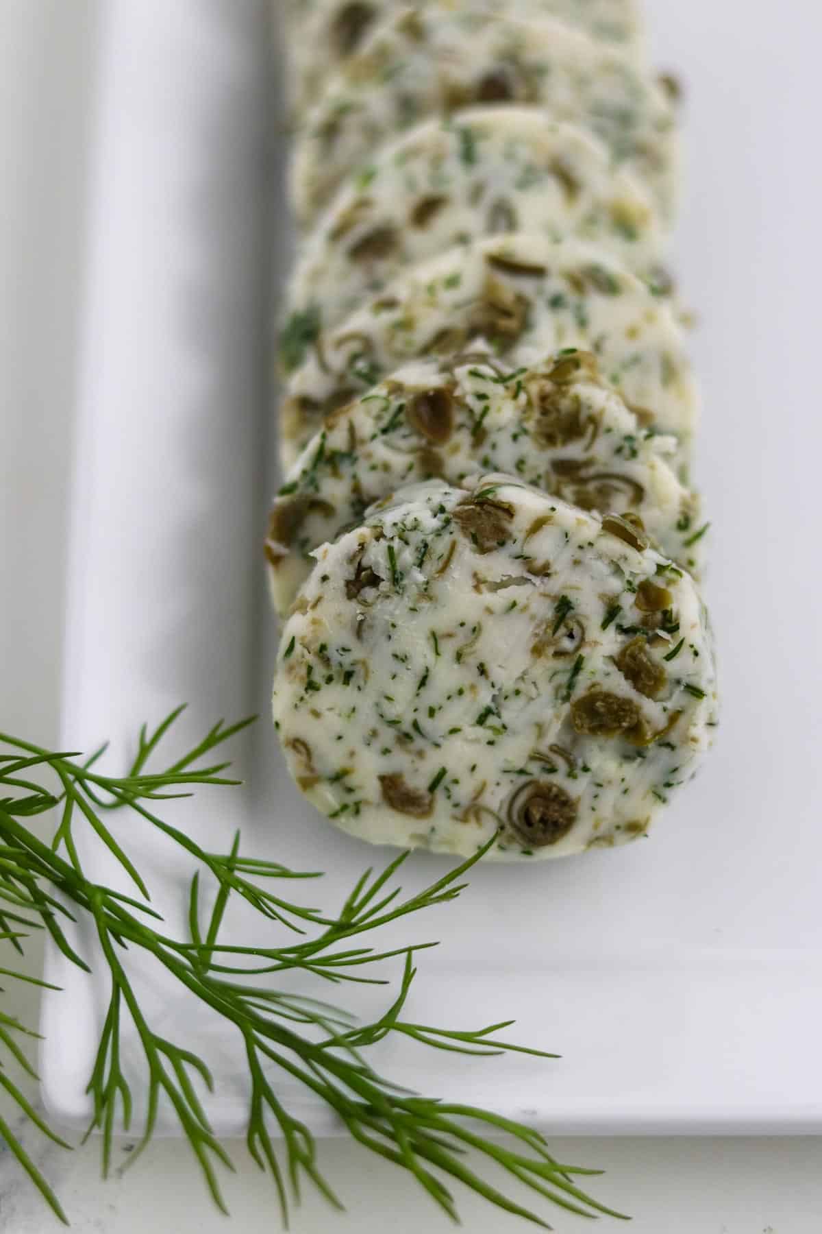 Rounds of Dill Caper Butter on a square plate next to a dill sprig.