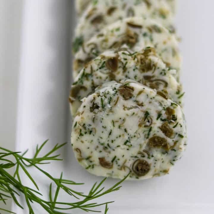 Close up of round of dill caper butter on a plate next to a sprig of dill.
