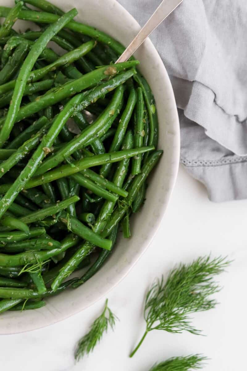 Green beans in a bowl with a spoon next to dill sprigs and a napkin.