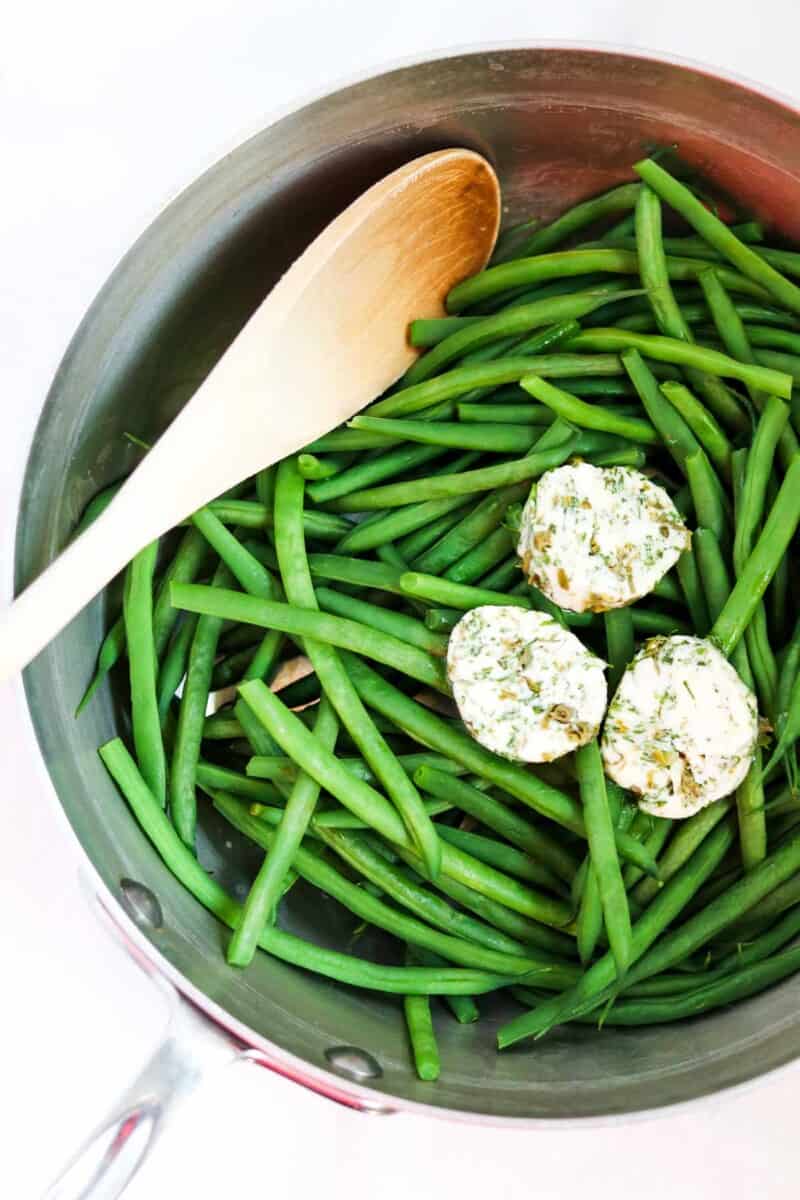 Green beans in a saucepan with Dill Caper Butter and a wooden spoon.