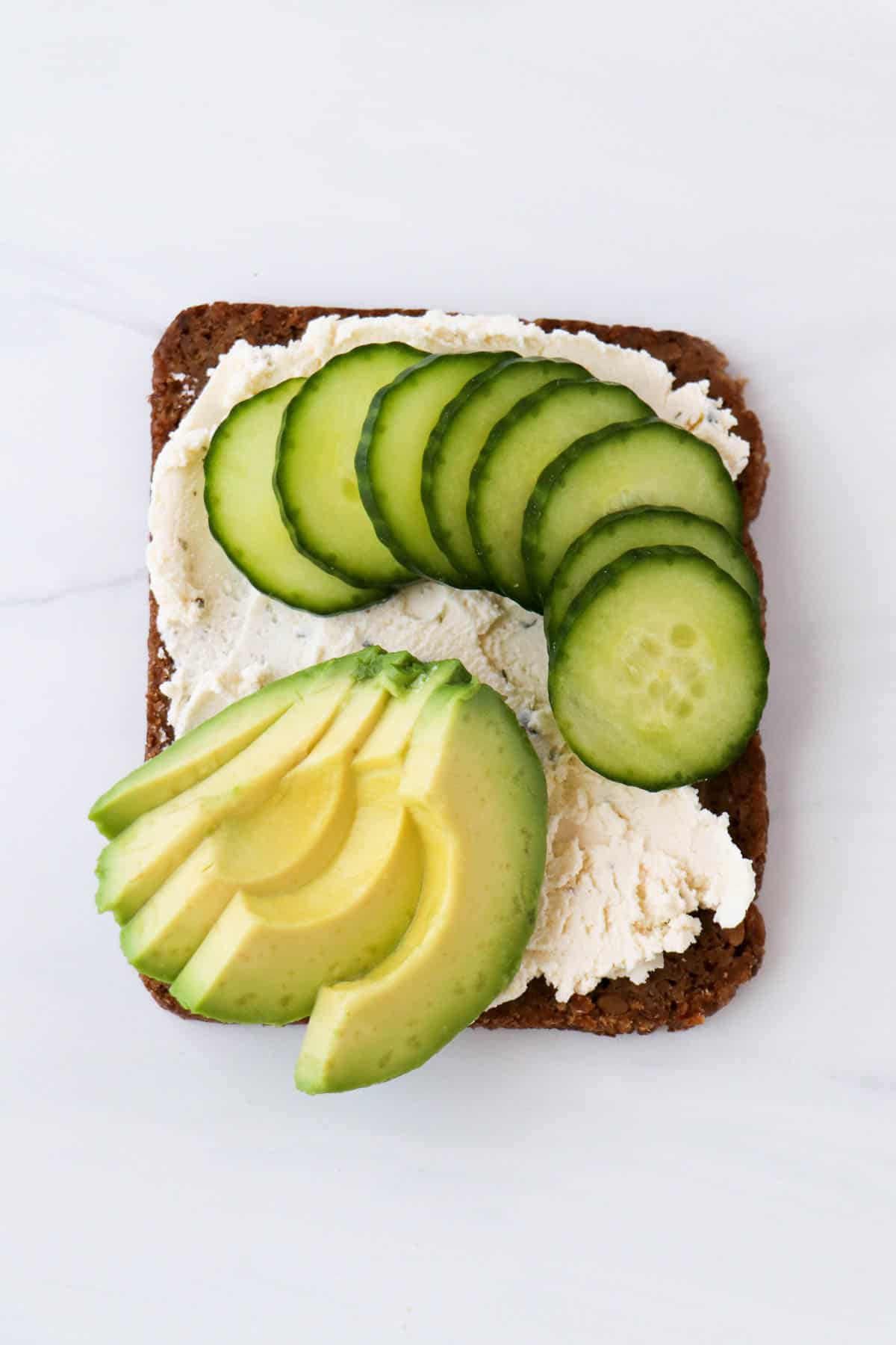 Sliced cucumbers and avocado on top of rye bread spread with soft cheese.