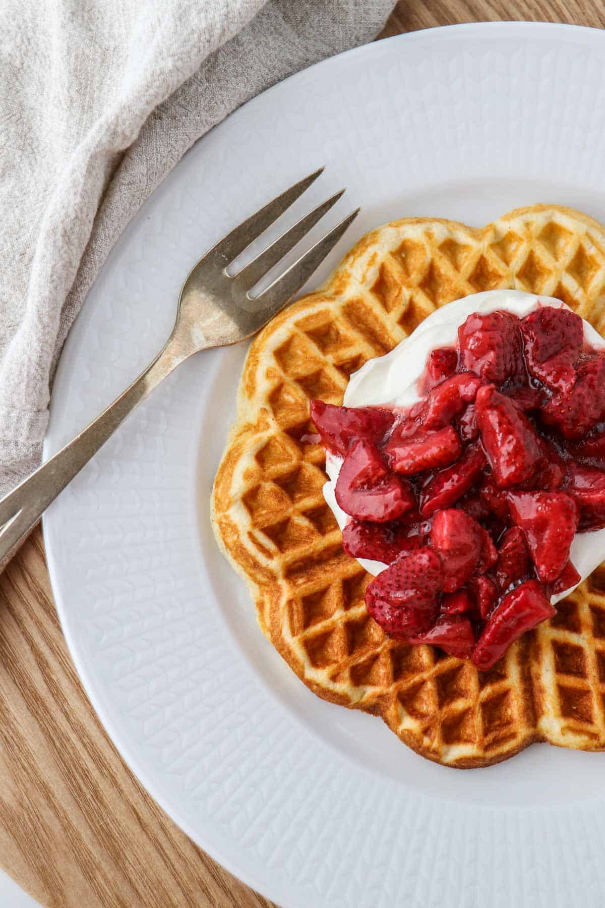 Heart shaped waffle on a plate topped with whipped cream and roasted strawberries.