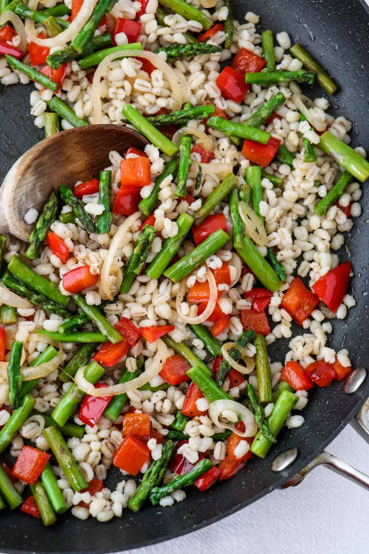 Red pepper, asparagus, shallot and barley in a skillet with a wooden spoon.
