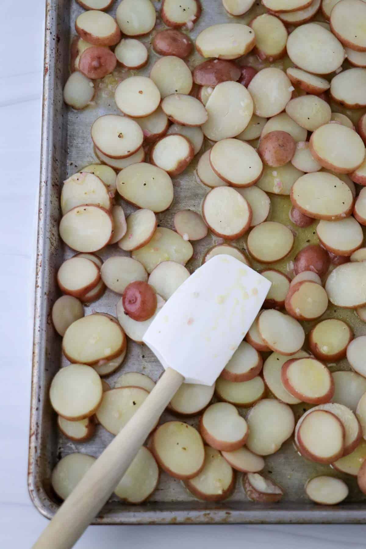 Sliced red potatoes on a baking sheet with a rubber spatula.