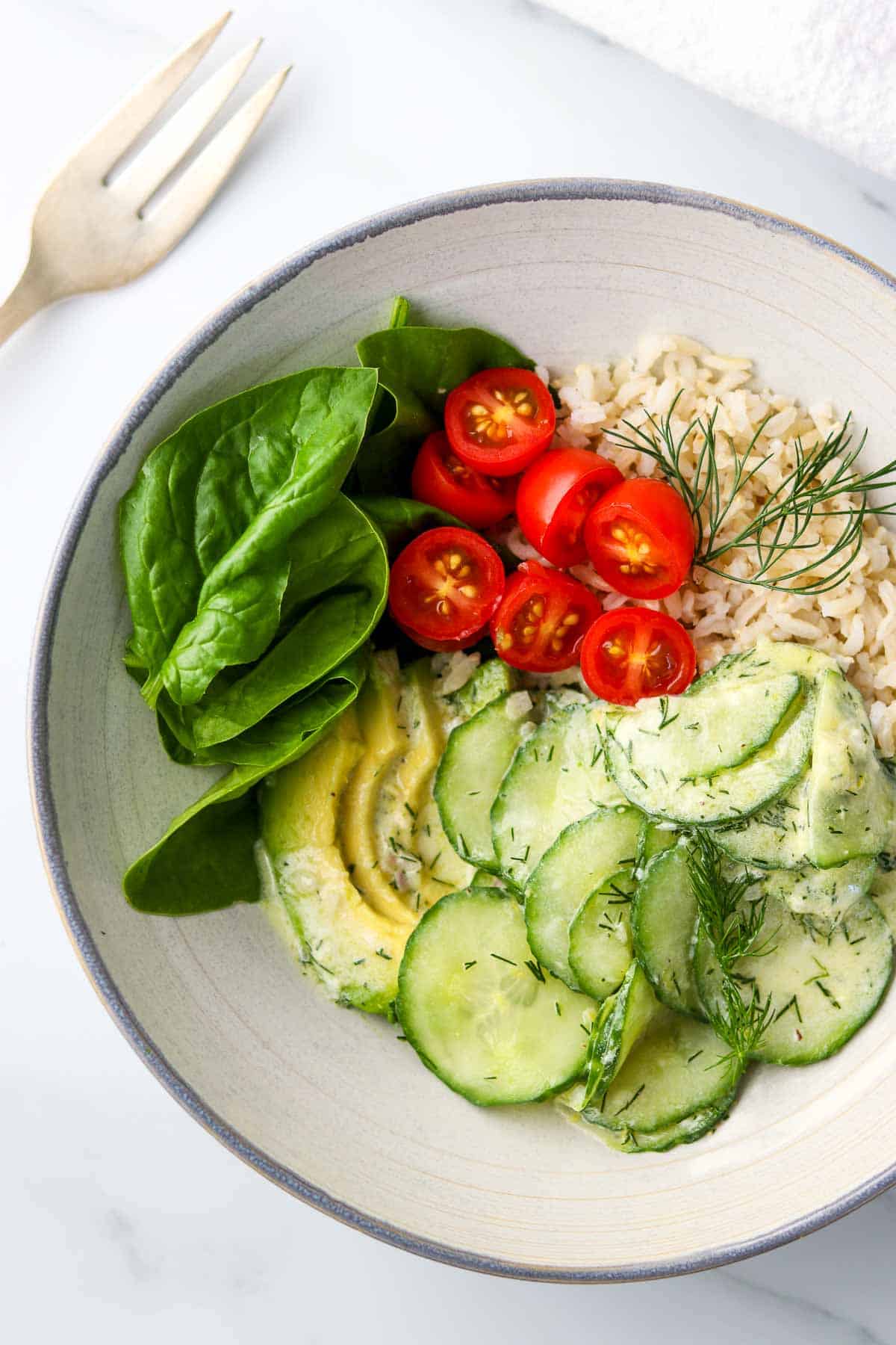 Cucumber salad, avocado, spinach, cherry tomatoes and rice in a bowl next to a fork.