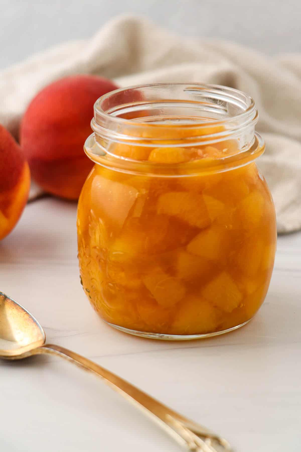Peach compote in a jar next to a spoon and fresh peaches.