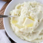 Overhead shot of mashed potatoes in a white bowl topped with butter with a spoon.