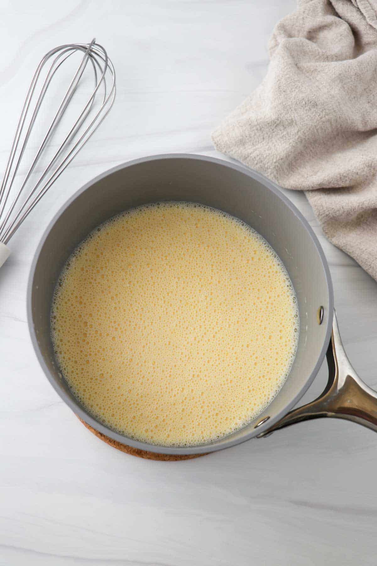 Vanilla sauce in a saucepan next to a whisk and a towel.