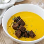 Close up of a bowl of butternut squash soup topped with rye croutons and herbs.