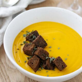 Close up of a bowl of butternut squash soup topped with rye croutons and herbs.