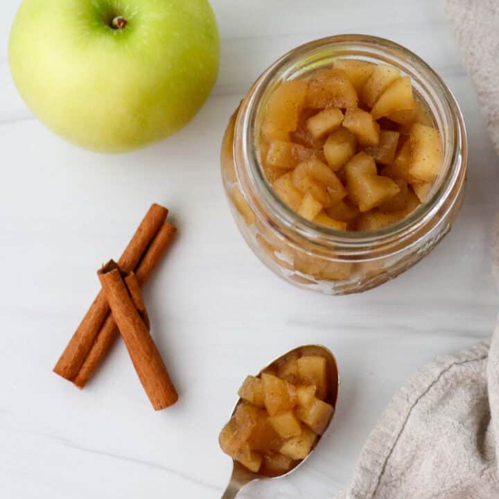 Jar of apple compote next to an apple, cinnamon sticks and a spoonful of compote.
