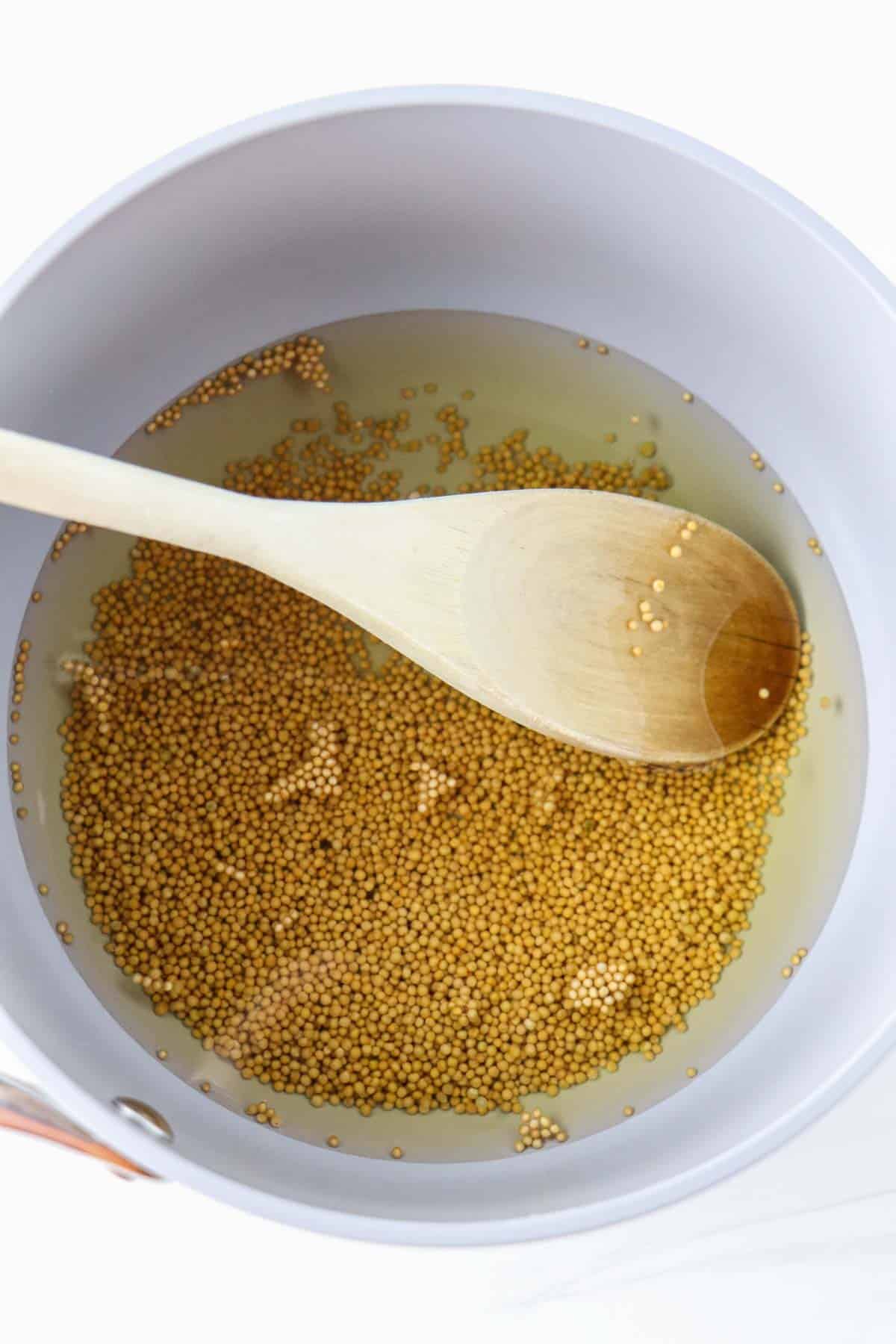 Mustard seeds in a pan with water and a wooden spoon.