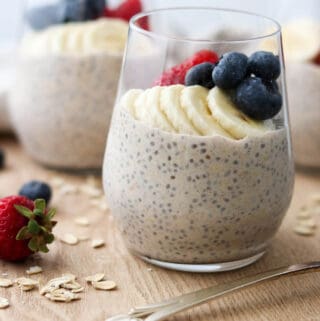 Close up of a glass of overnight oats topped wit bananas, blueberries and strawberries next to raw oats and a spoon.