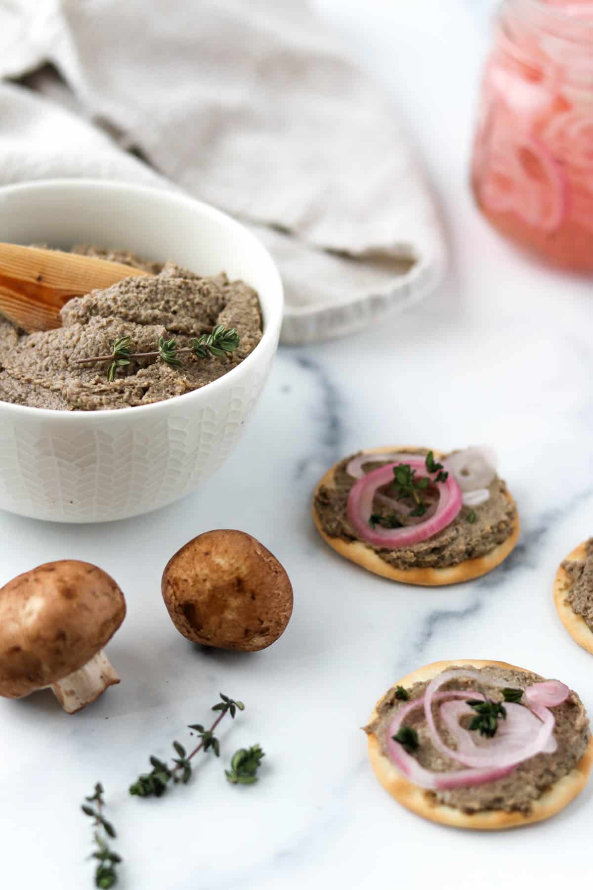 Vegetarian Mushroom Pâté in a bowl next to mushrooms and crackers topped with pate and pickled shallots.