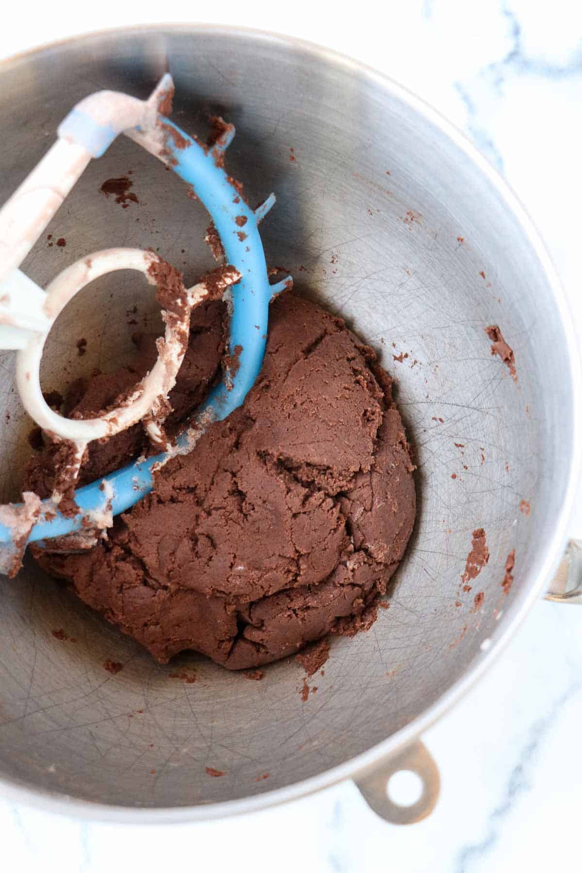 Chocolate cookie dough in a metal bowl with a blue paddle attachment for a mixer.