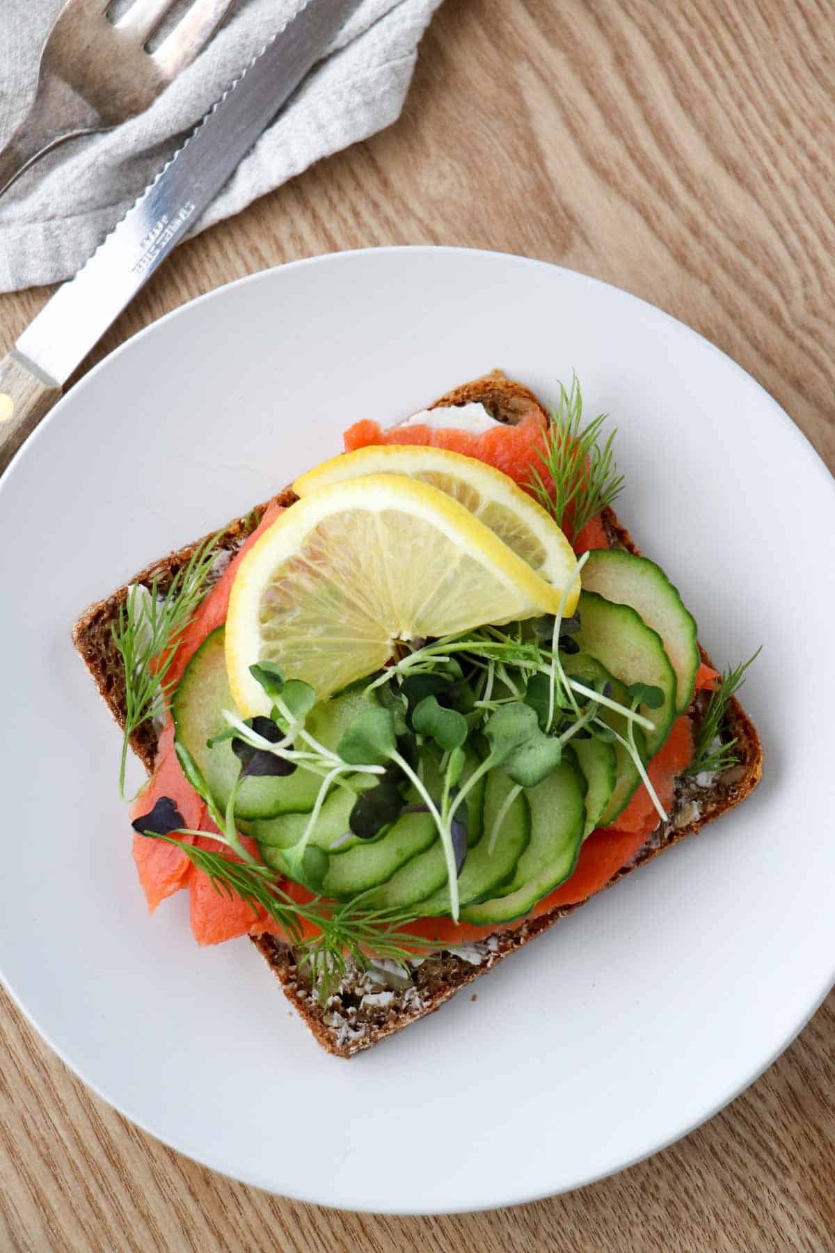 Smoked salmon smørrebrød on a white plate next to a knife and fork.