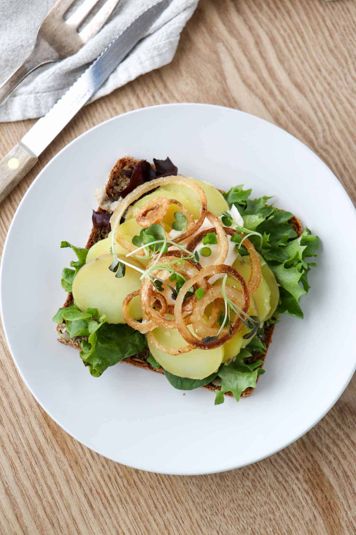 Potato smørrebrød on a white plate next to a knife and fork.