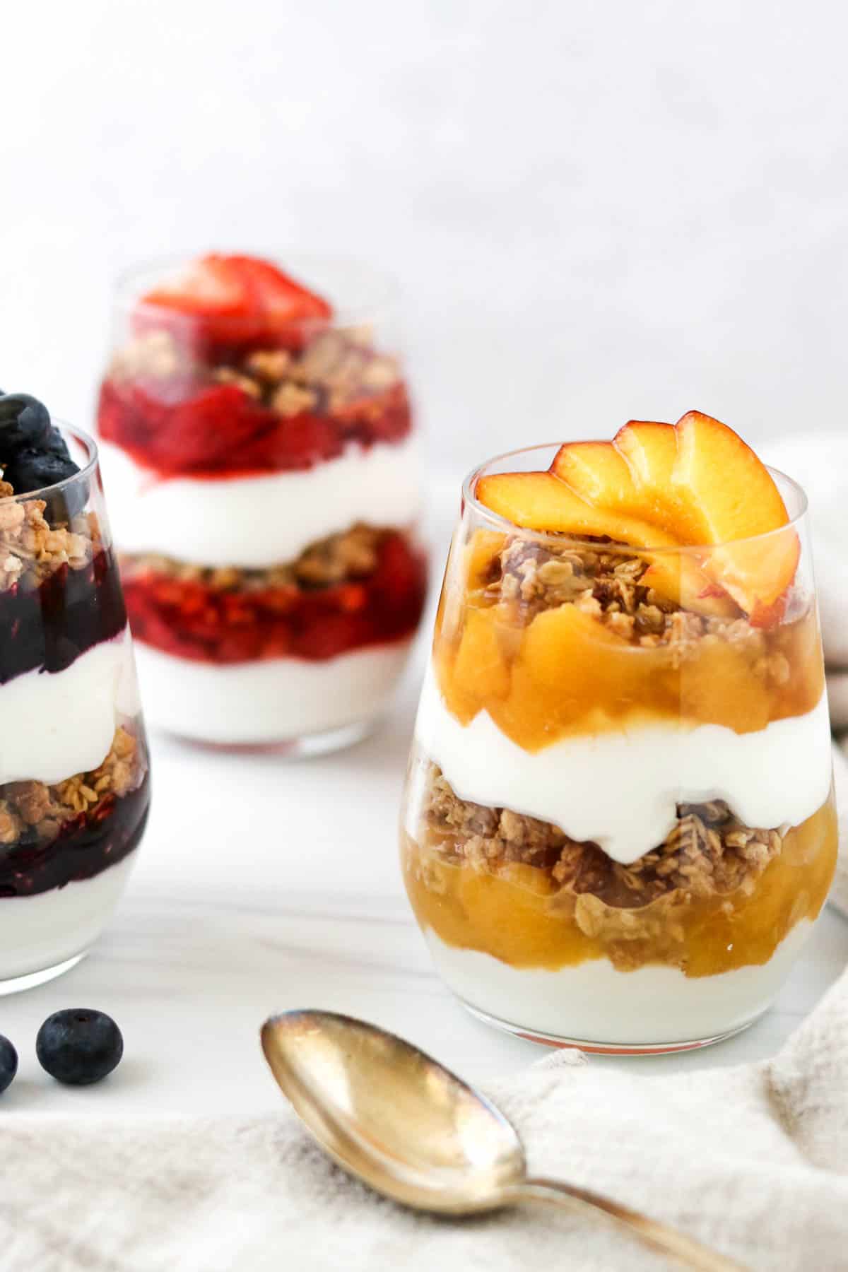 Peach, blueberry and strawberry yogurt parfaits next to a spoon and fresh blueberries.