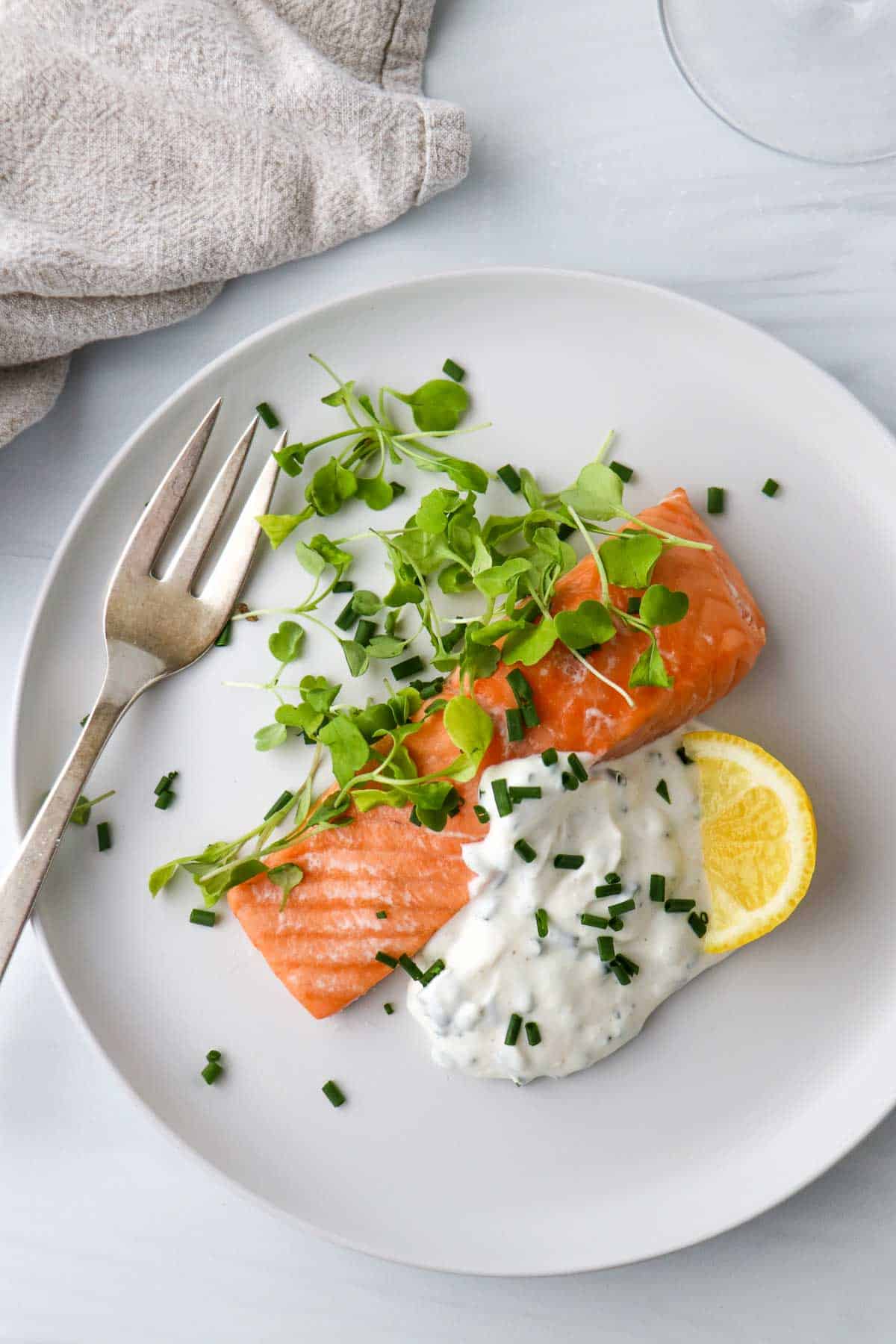 Overhead view of salmon, greens, horseradish yogurt sauce and a lemon wedge on a white plate next to a fork.