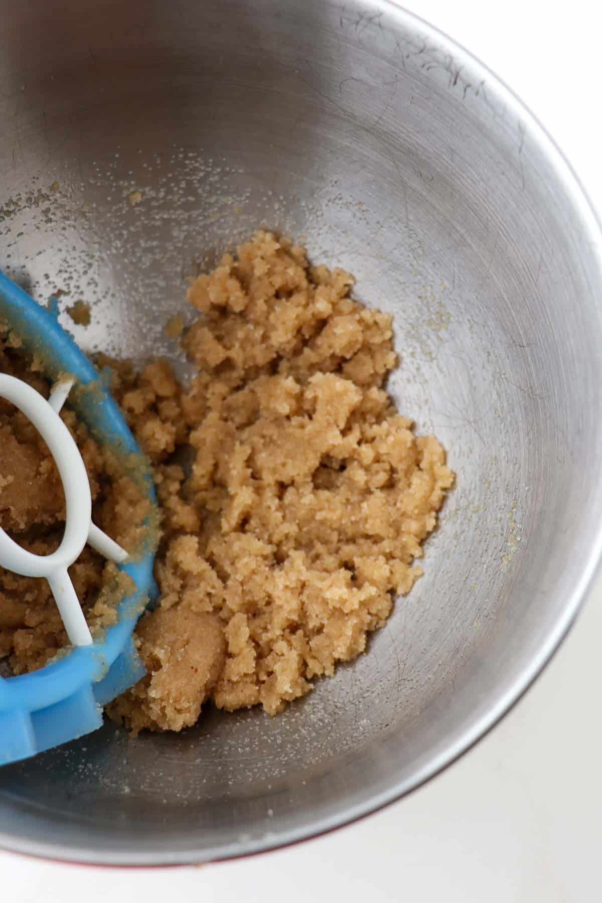 Brown sugar, sugar and oil mixed together in a metal mixing bowl with a mixer paddle attachment.