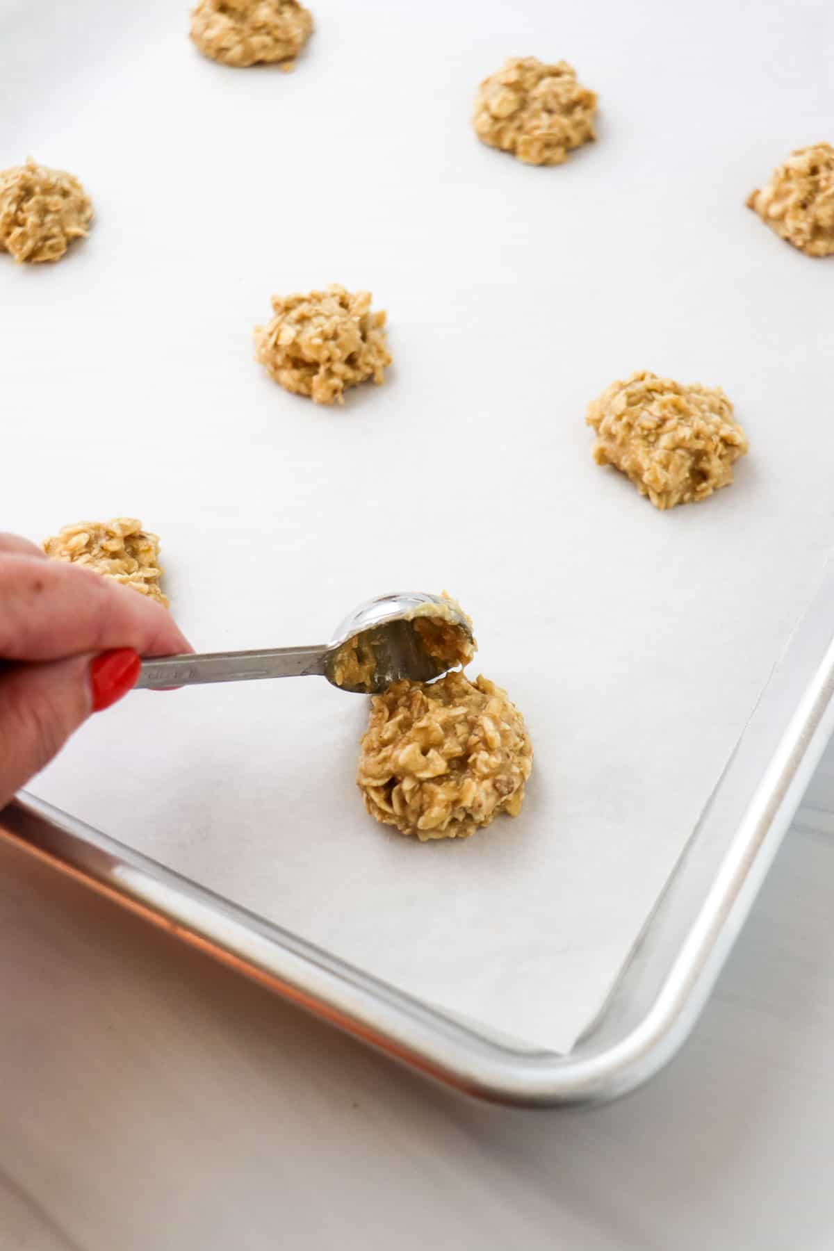 Person dropping spoonfuls of oatmeal cookie dough on a parchment lined baking sheet.