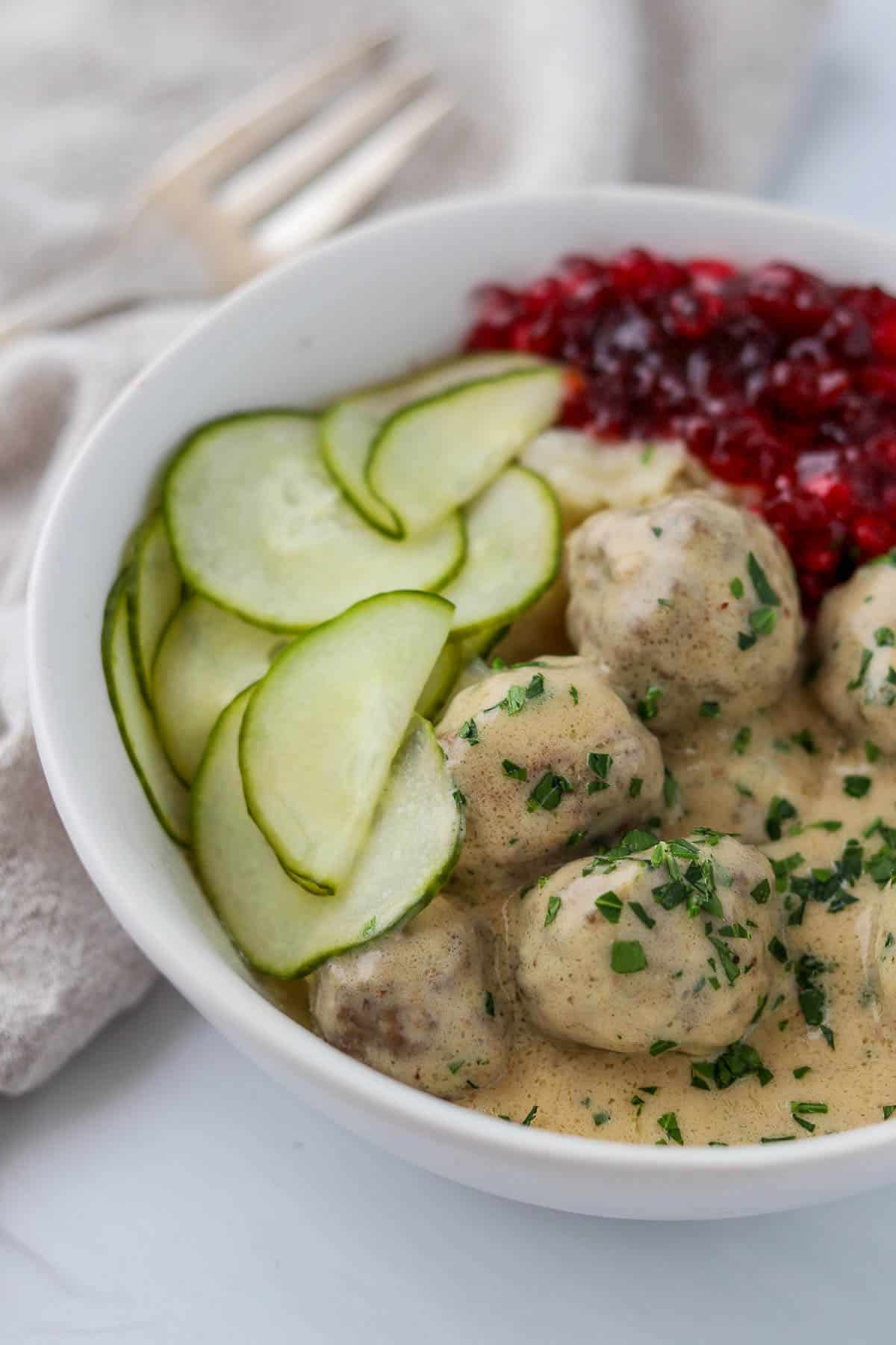 Swedish meatballs in a bowl with cucumbers and lingonberries.