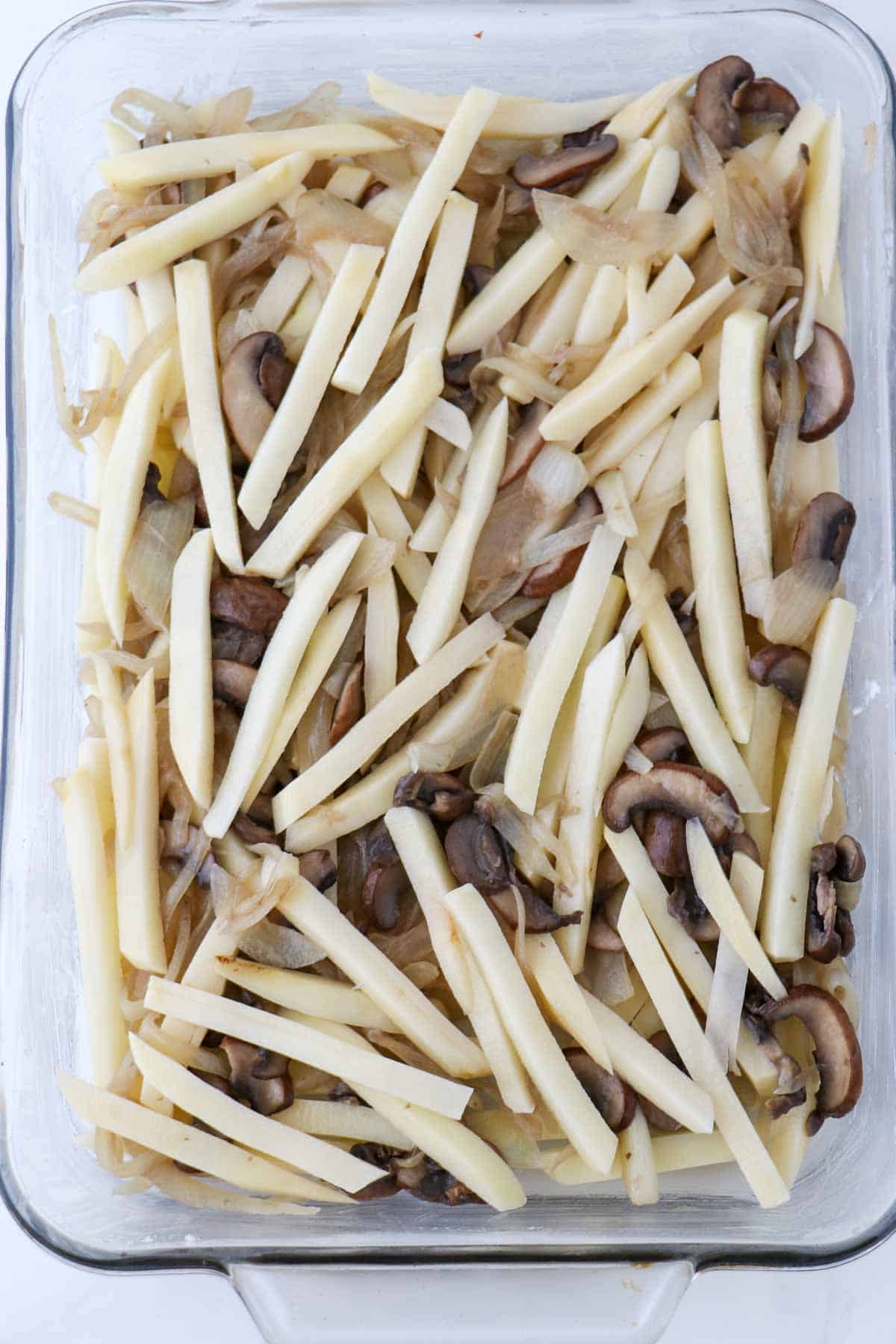 Sliced potatoes, onions and mushrooms layered together in a glass baking dish.