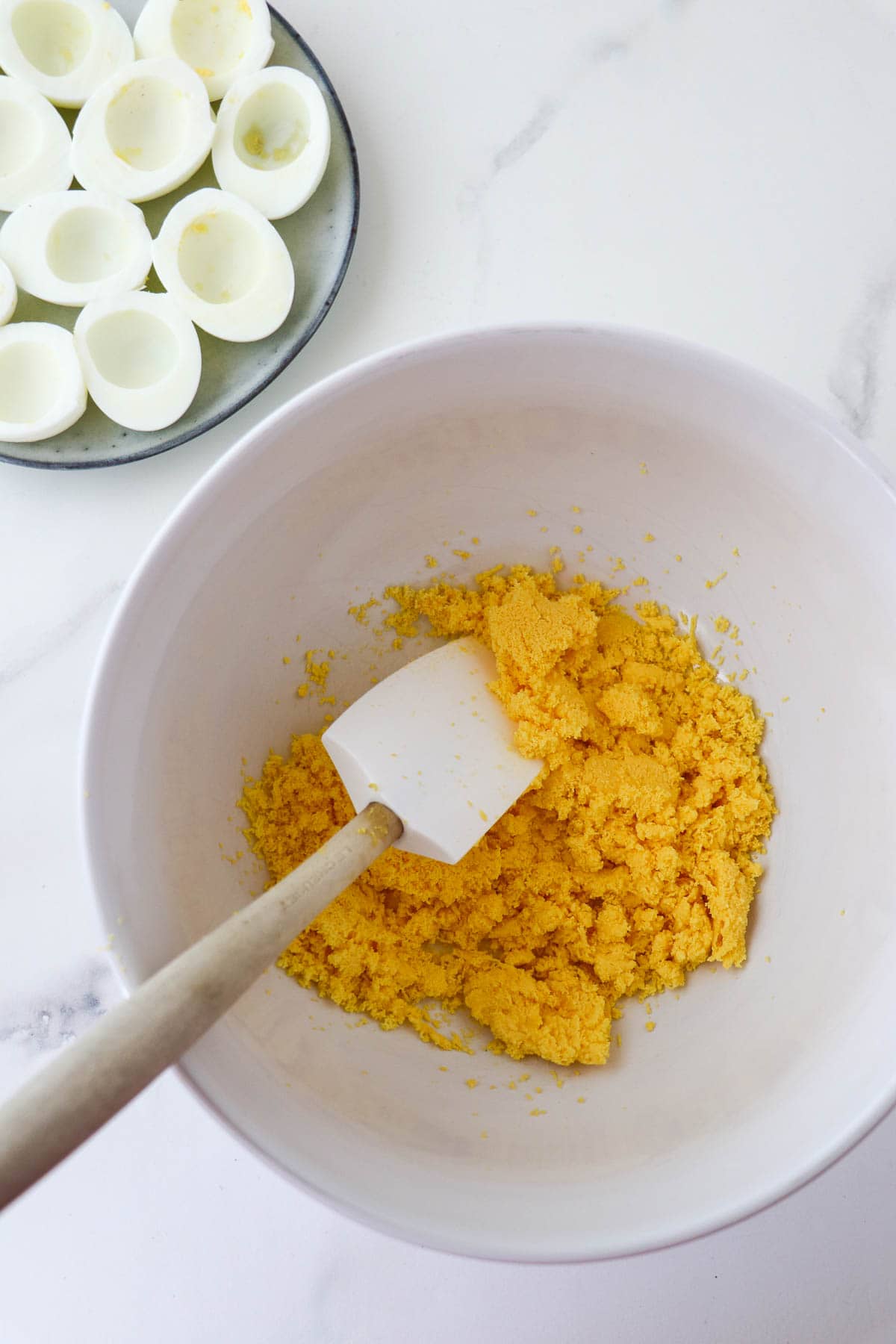 Mashed up egg yolks in a bowl with a rubber spatula next to egg white halves.
