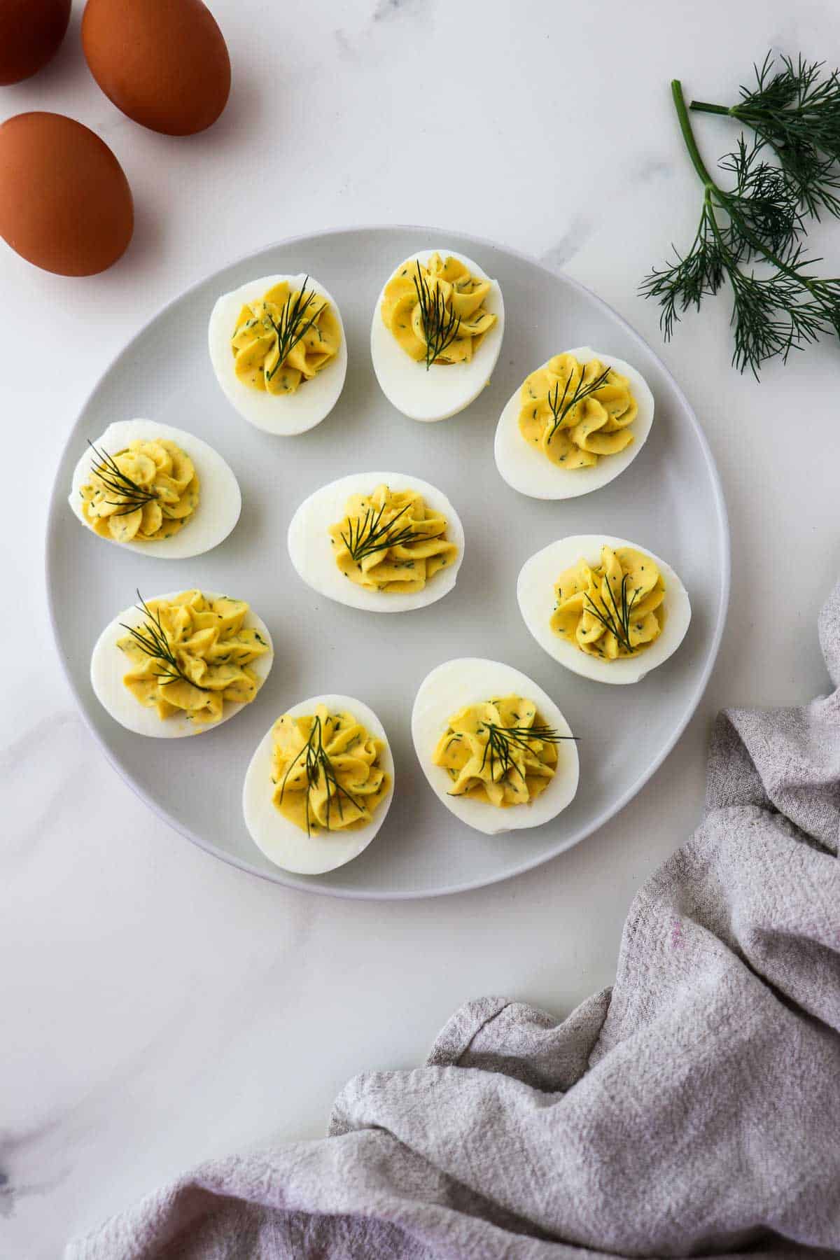 Deviled Eggs with Dill on a plate next to eggs, dill sprig and kitchen towel.